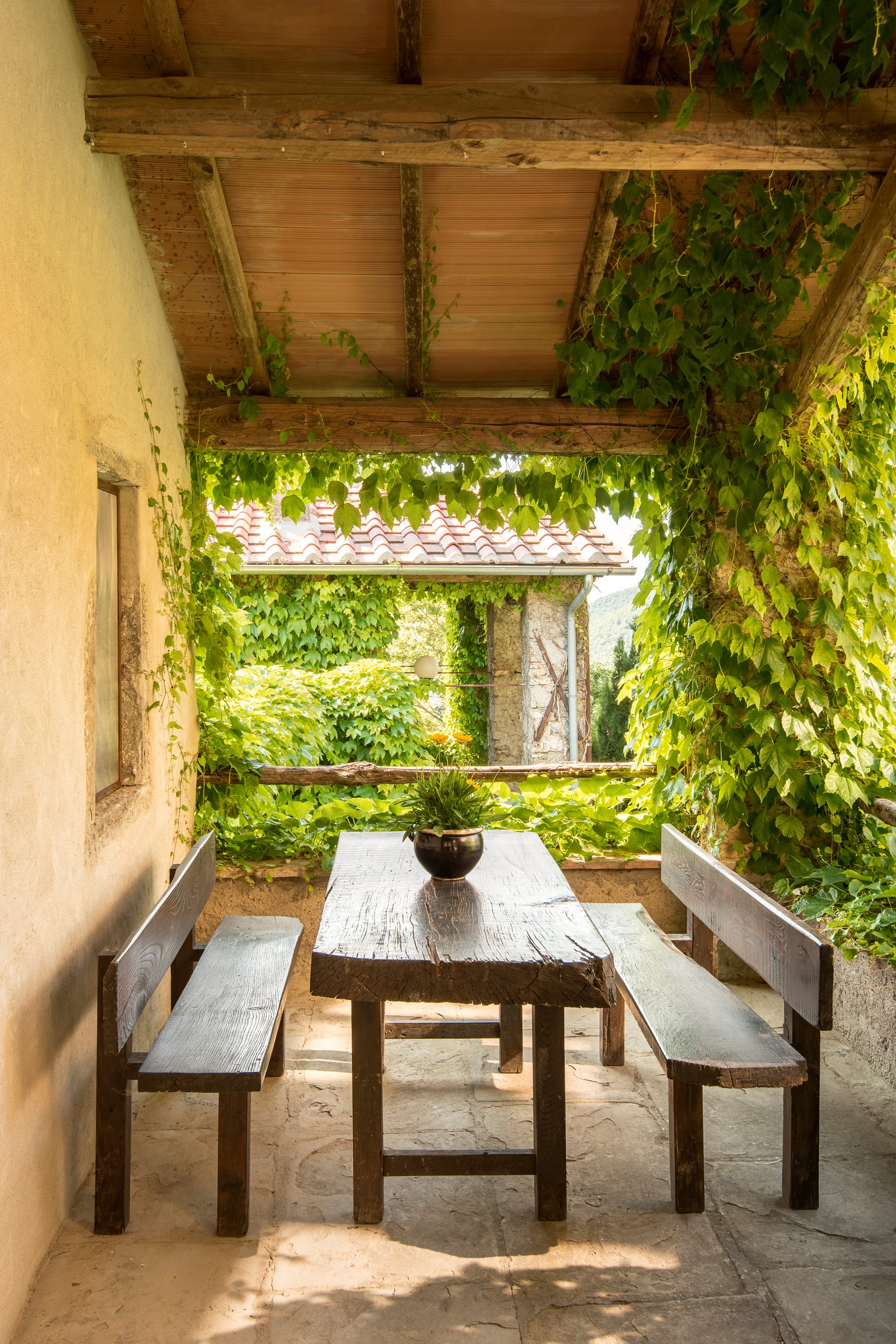 Wooden table and benches in a covered stone loggia, framed by climbing plants