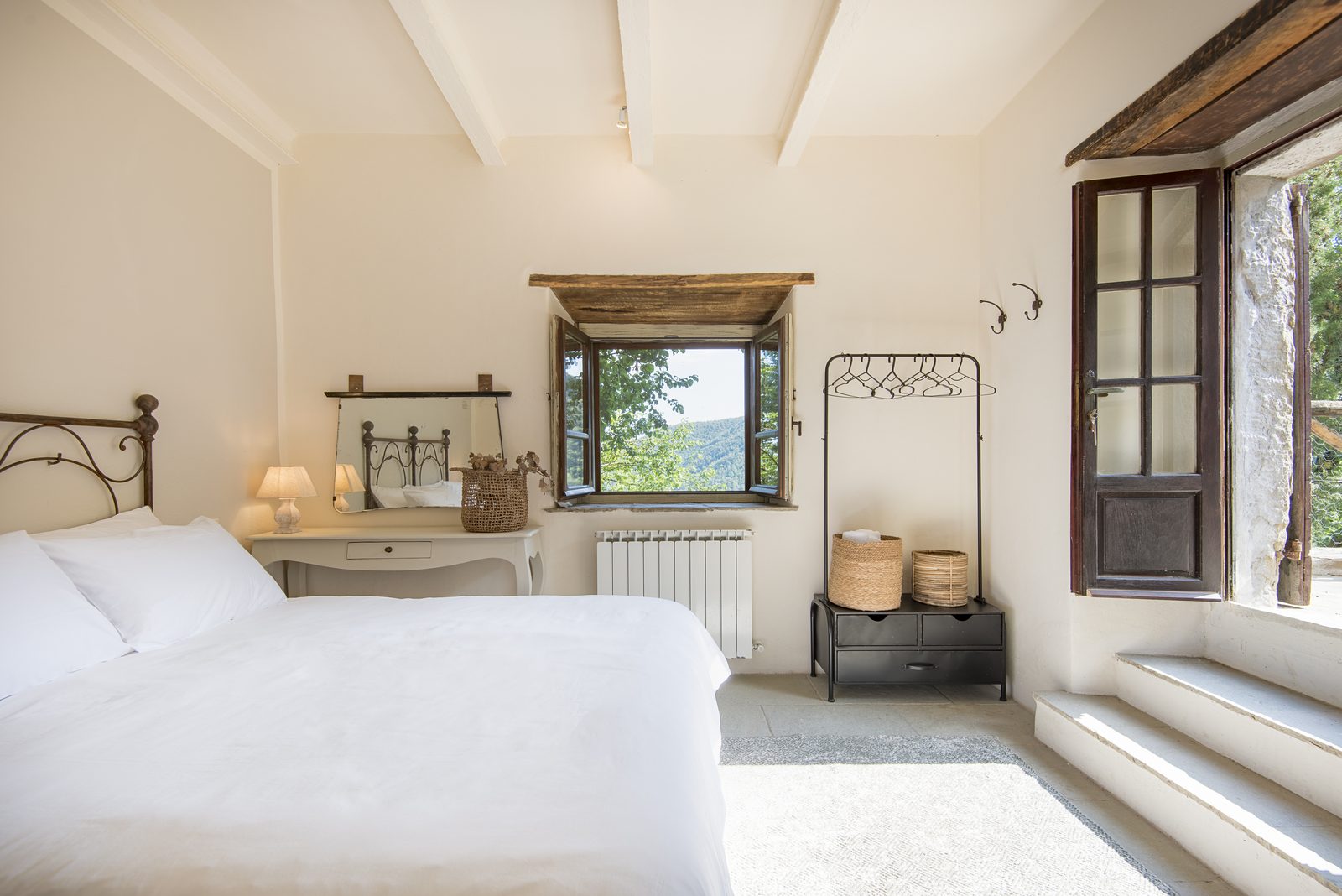 Guest house suite: a bright bedroom with a white bed, an iron headboard, wooden beams, a small window onto the valley and stone steps up to an open door