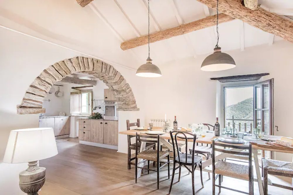 Main house: open-plan dining room with a long wooden table under two grey pendant lamps, a stone arch into the kitchen and a window onto the valley