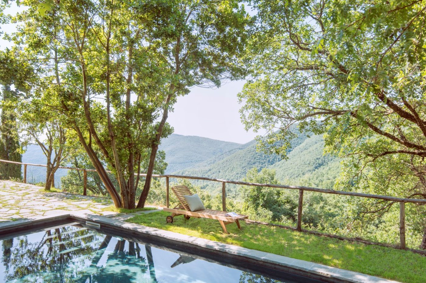 The edge of the saltwater pool, with a wooden sun lounger on the grass and trees framing a view over the wooded Val di Pierle