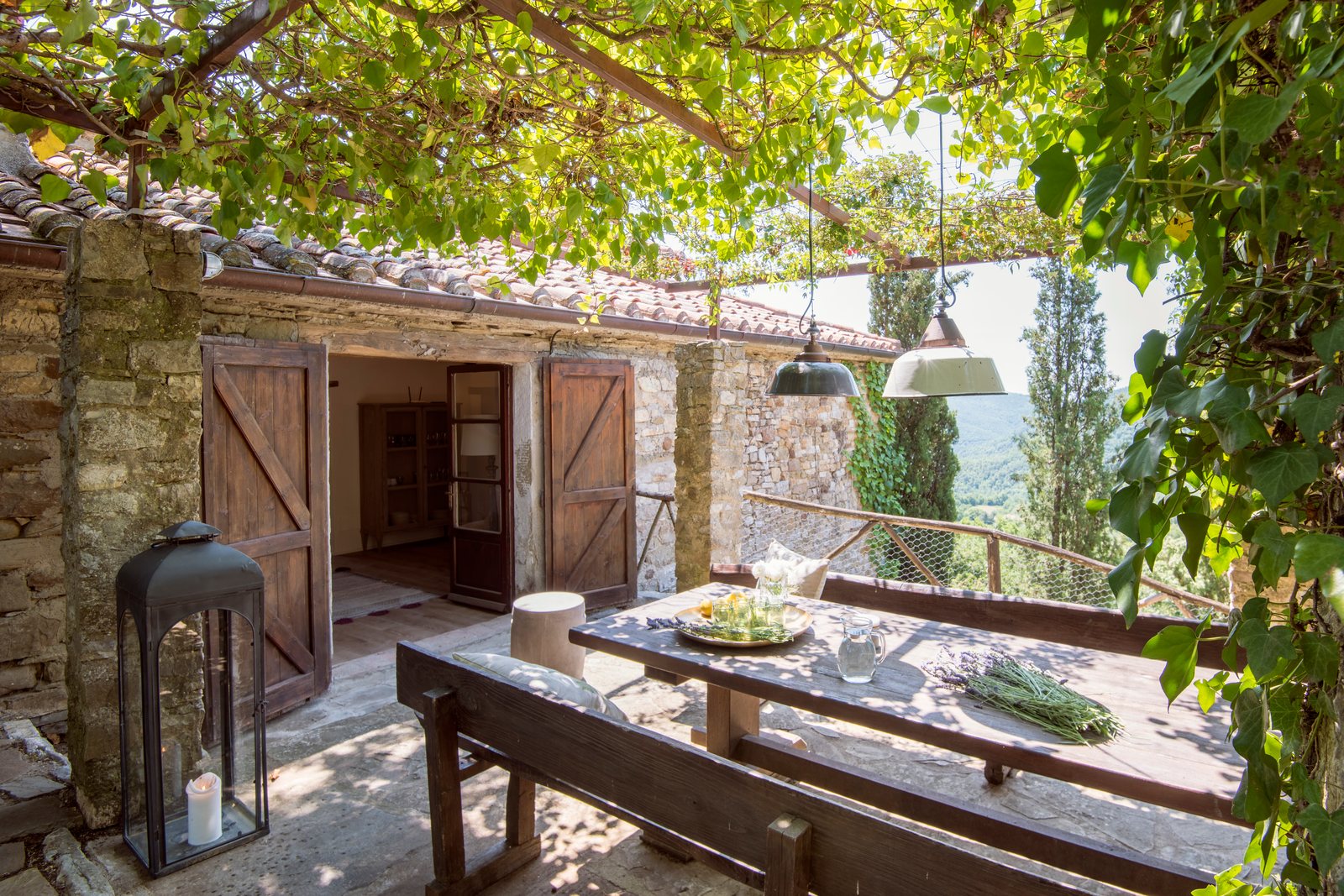 Stone terrace under a vine pergola, with a long wooden table beside open barn doors
